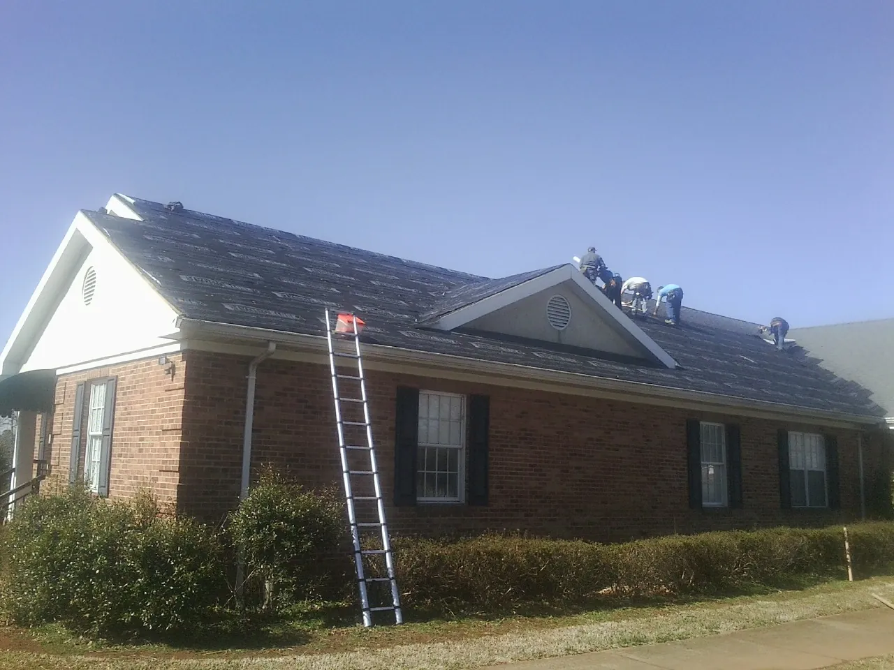 Crew tearing shingles off a brick ranch roof with a ladder set — Metal Roof Repair work by our crew in Drewryville