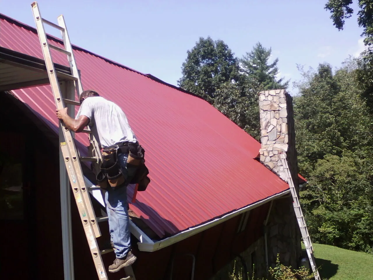 Roofer on a ladder beside a red metal roof and stone chimney — Soffit & Fascia Repair by Roofing Brothers in Drewryville